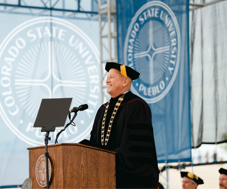 A man stands at a podium in graduation regalia.