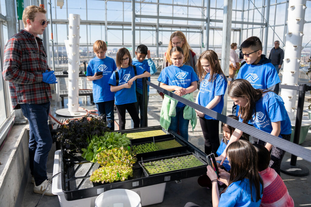A group of kids in matching shirts gather around a speaker in a greenhouse.