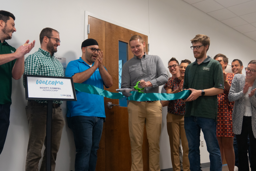 A group of people cut a blue and green ribbon in front of a door.