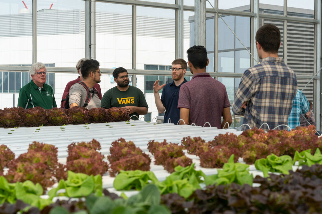 A group gathers around a speaker in a greenhouse with a table of plants in the foreground.