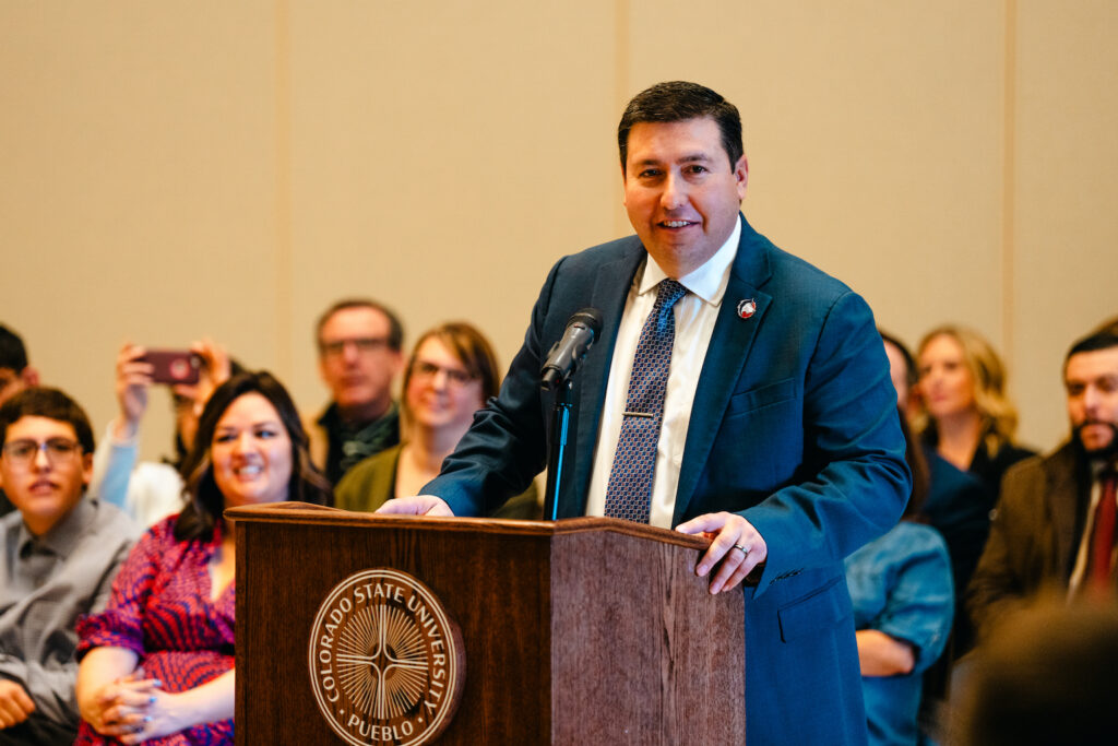 Man in a blue suit with a CSU Pueblo lapel pin stands at a podium.
