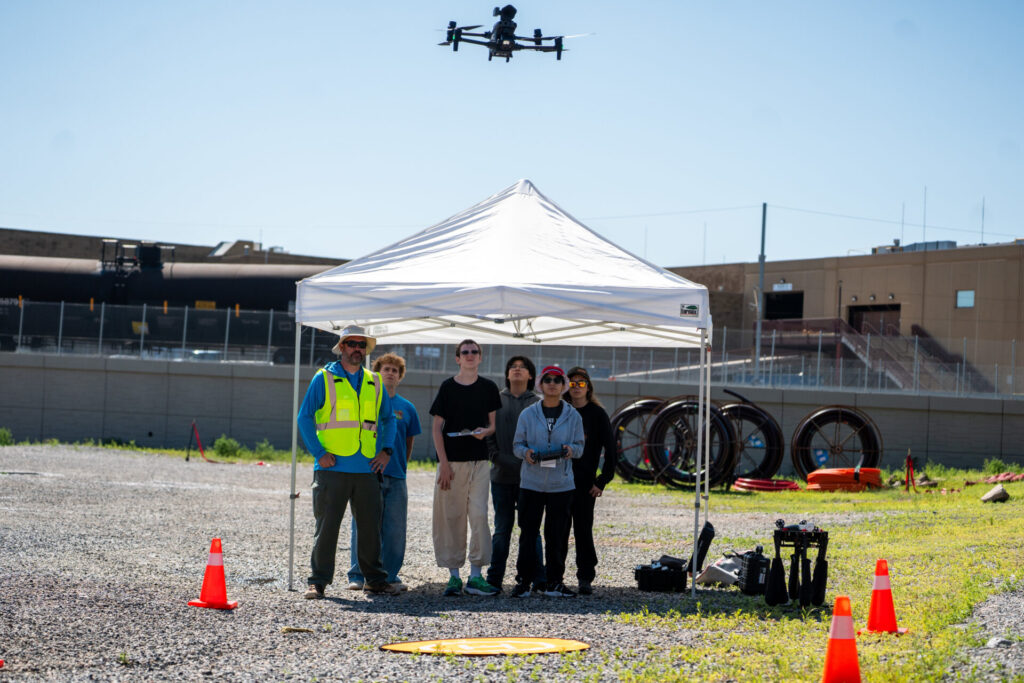 A group of students stand under a white tent while flying a drone.