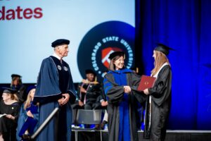 Becky Takeda-Tinker, in academic regalia, hands a diploma to a graduating student on stage.