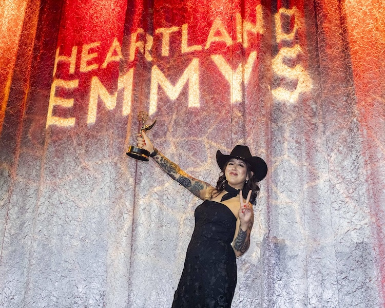 A woman in a black dress and black cowboy hat holds up an Emmy statue on stage in front of a curtain with the words "Heartland Emmys."